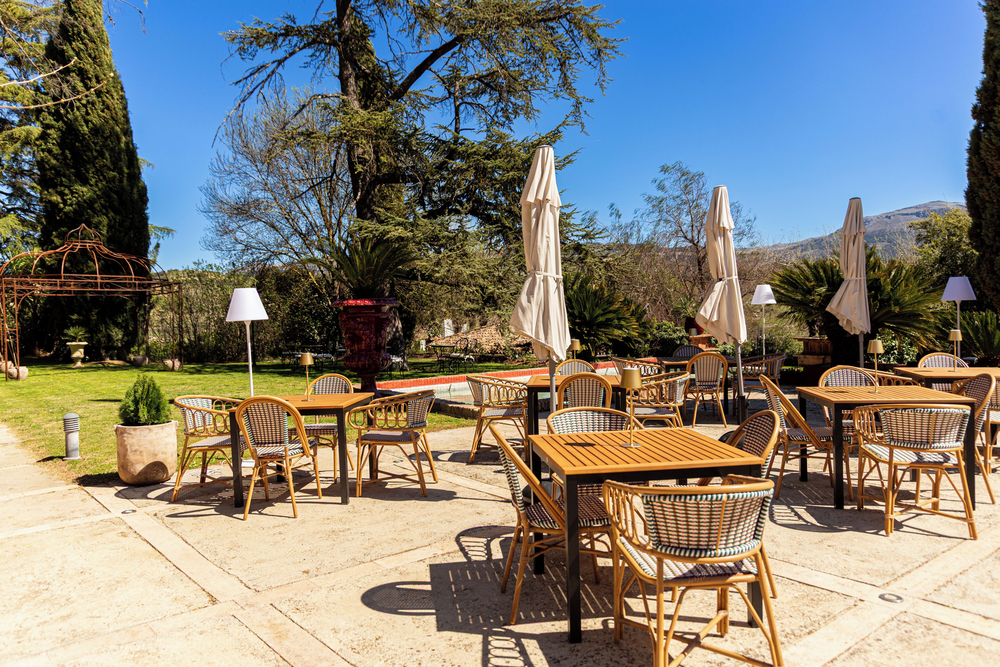 An outdoor patio with wicker furniture and umbrellas, surrounded by trees and a mountain in the background.