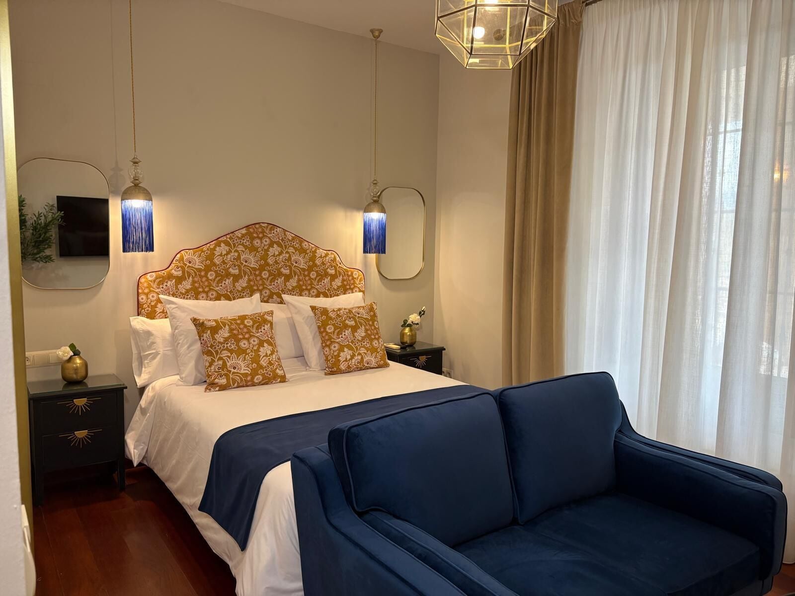 A hotel room with a bed, a blue sofa, a nightstand with a lamp, and a mirror.