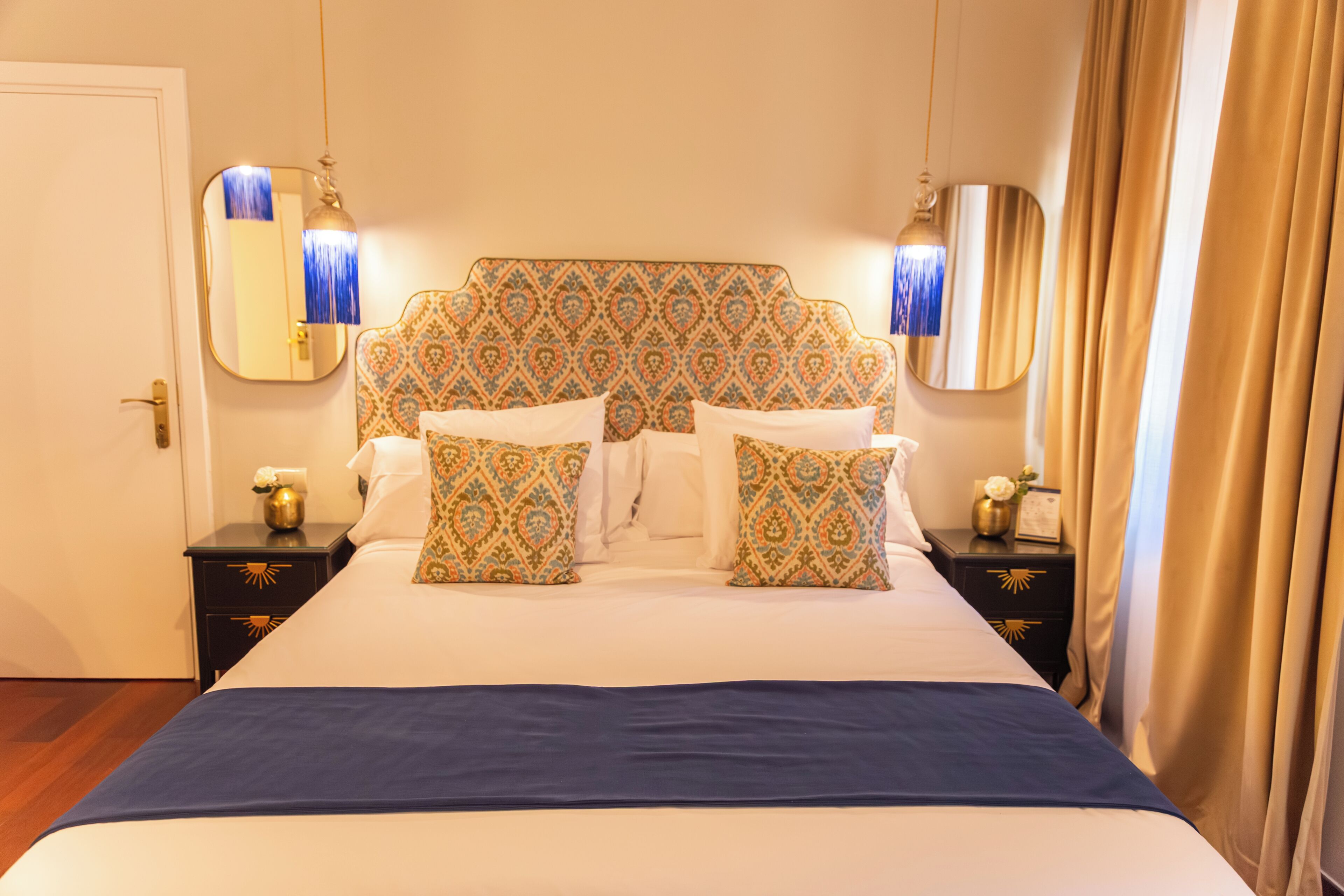 A hotel room with a large bed, patterned headboard, bedside tables with lamps, and a mirror.