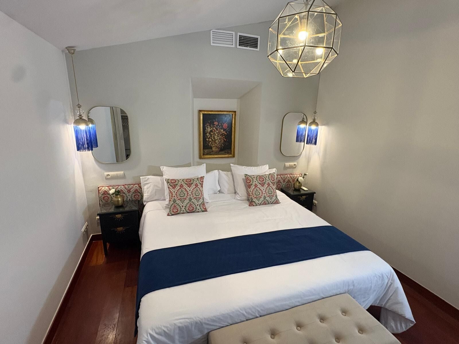Une chambre d’hôtel avec un grand lit, deux tables de chevet, un climatiseur fixé au mur et un luminaire suspendu au design géométrique.