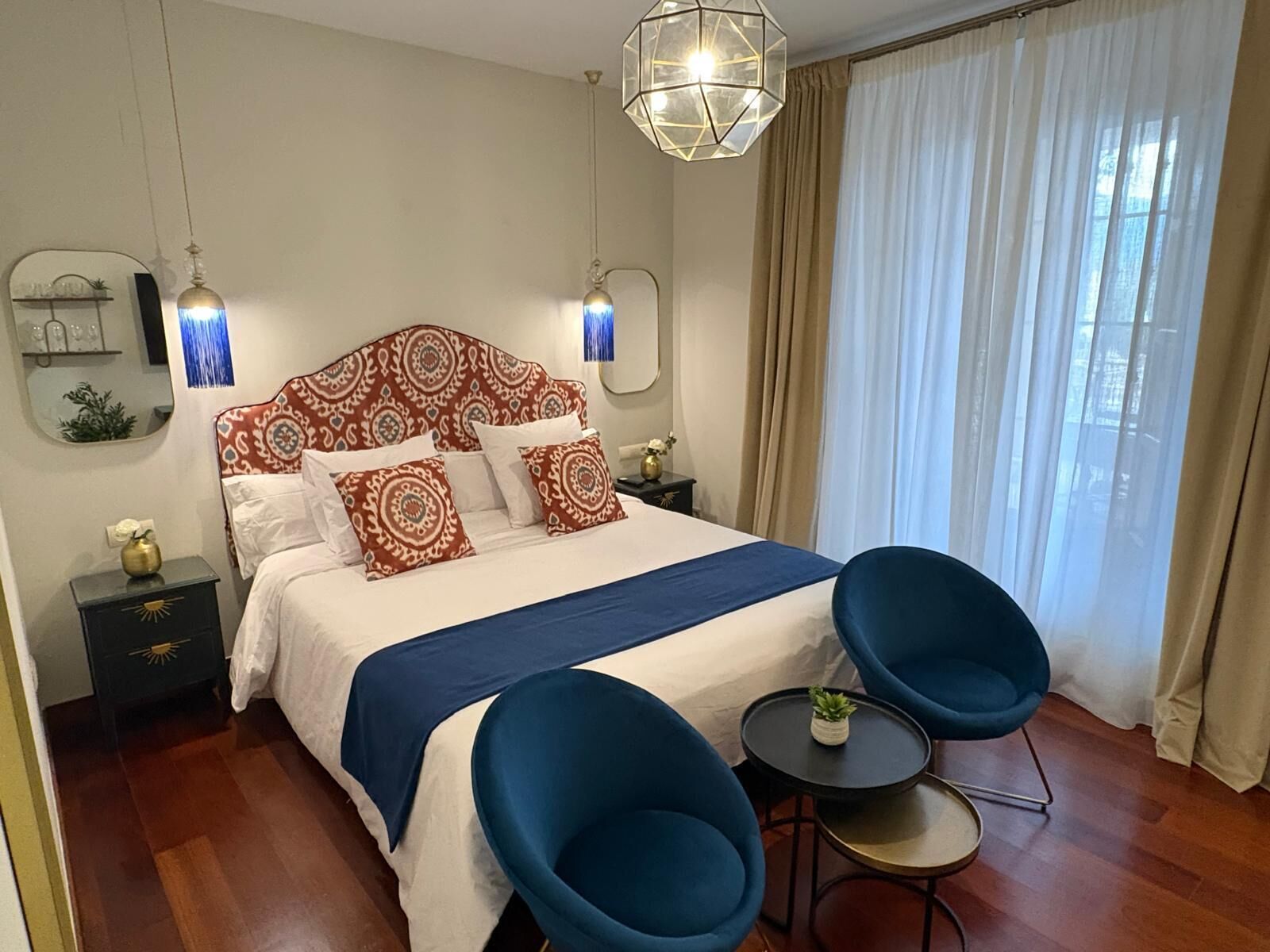 A modern hotel room with a large bed, two blue chairs, a small table, and a decorative ceiling light.
