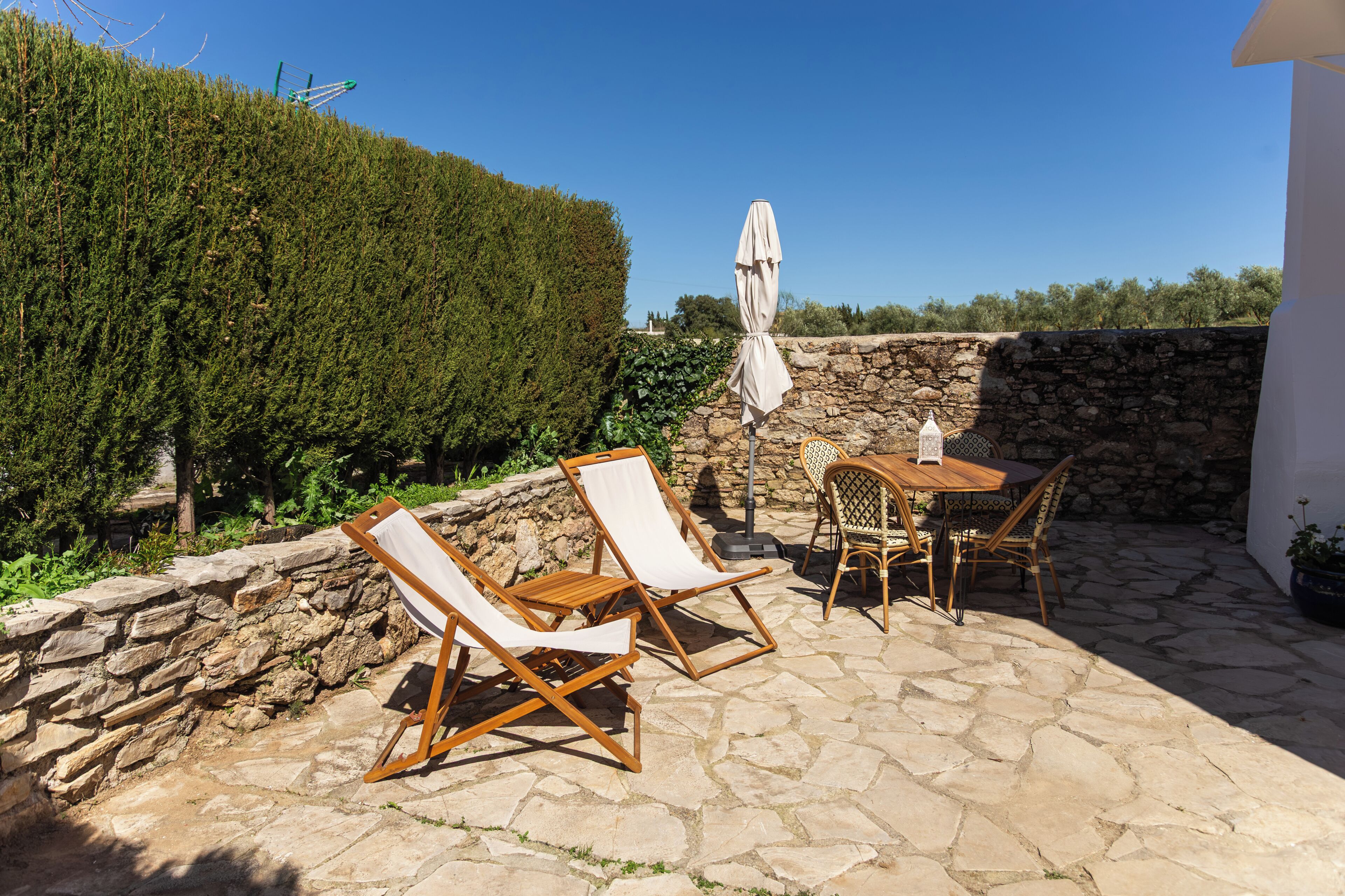 A patio with stone flooring, wooden lounge chairs, a table, and a white umbrella.