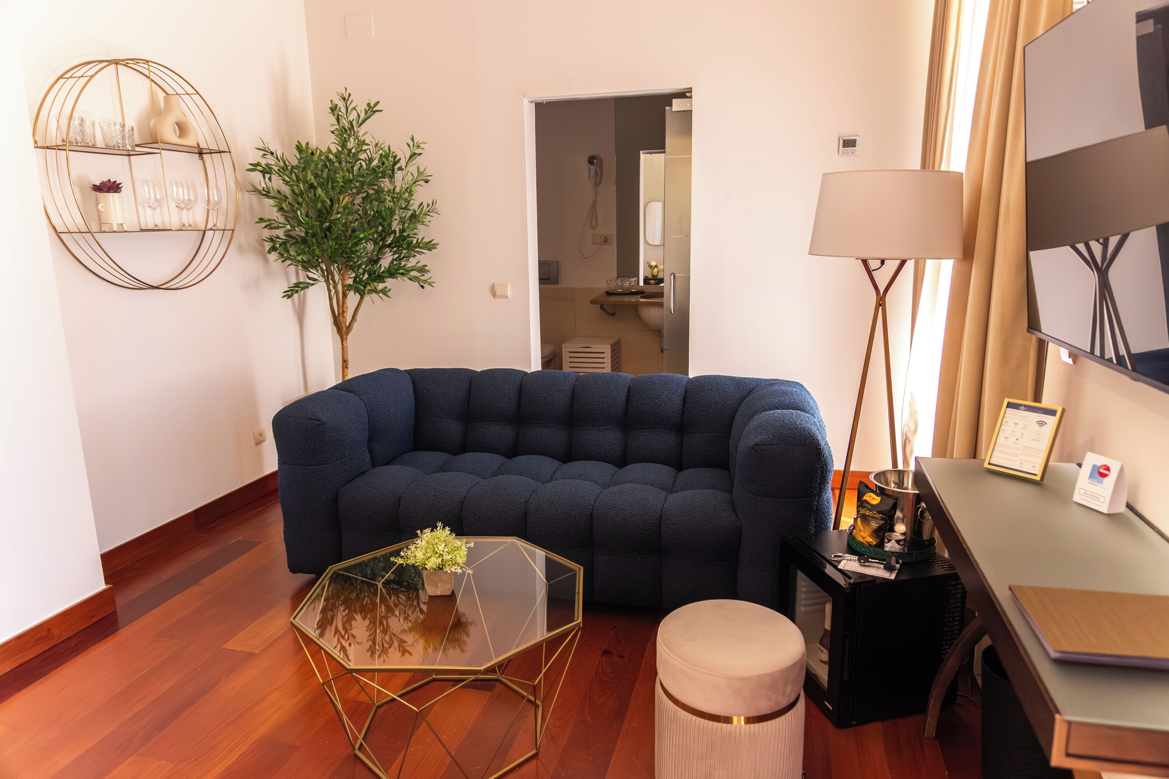 A living room with a dark blue sofa, a glass coffee table, a small round ottoman, a wall-mounted shelf with decorative items, a potted plant, and a lamp.