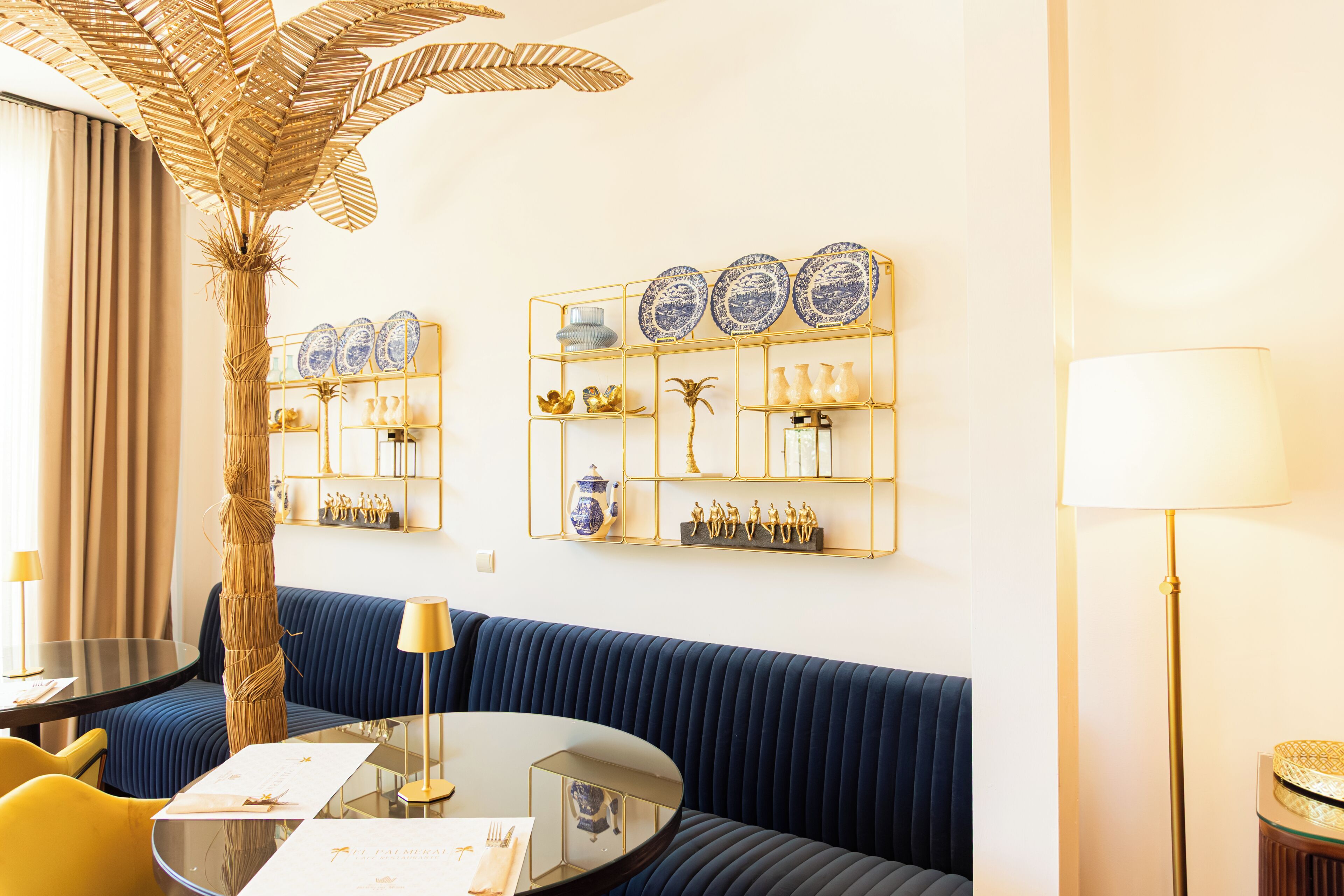 A room with a blue sofa, a round glass table, a gold-framed shelf with decorative items, a tall palm plant, and a floor lamp.