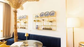 A room with a blue sofa, a round glass table, a gold-framed shelf with decorative items, a tall palm plant, and a floor lamp.