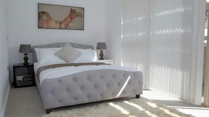 Un dormitorio con una cama grande, dos mesitas de noche con lámparas, un cuadro en la pared y vistas al exterior a través de cortinas translúcidas.