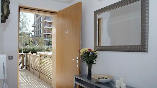 Un pasillo con una puerta de madera, un espejo, un jarrón con flores, un cuenco y una mesita.