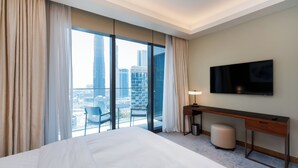Habitación de hotel con una cama grande, un escritorio, una silla, un televisor y vistas panorámicas de la ciudad a través de amplios ventanales.