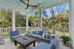 A covered outdoor seating area with a sofa, chairs, and a table, surrounded by white railings and a view of tropical vegetation. - New, Luxurious Coastal Gem! Private Pool, Game Room, Putt-Putt, Easy Walk to Beach, & More! (Isle Of Palms)