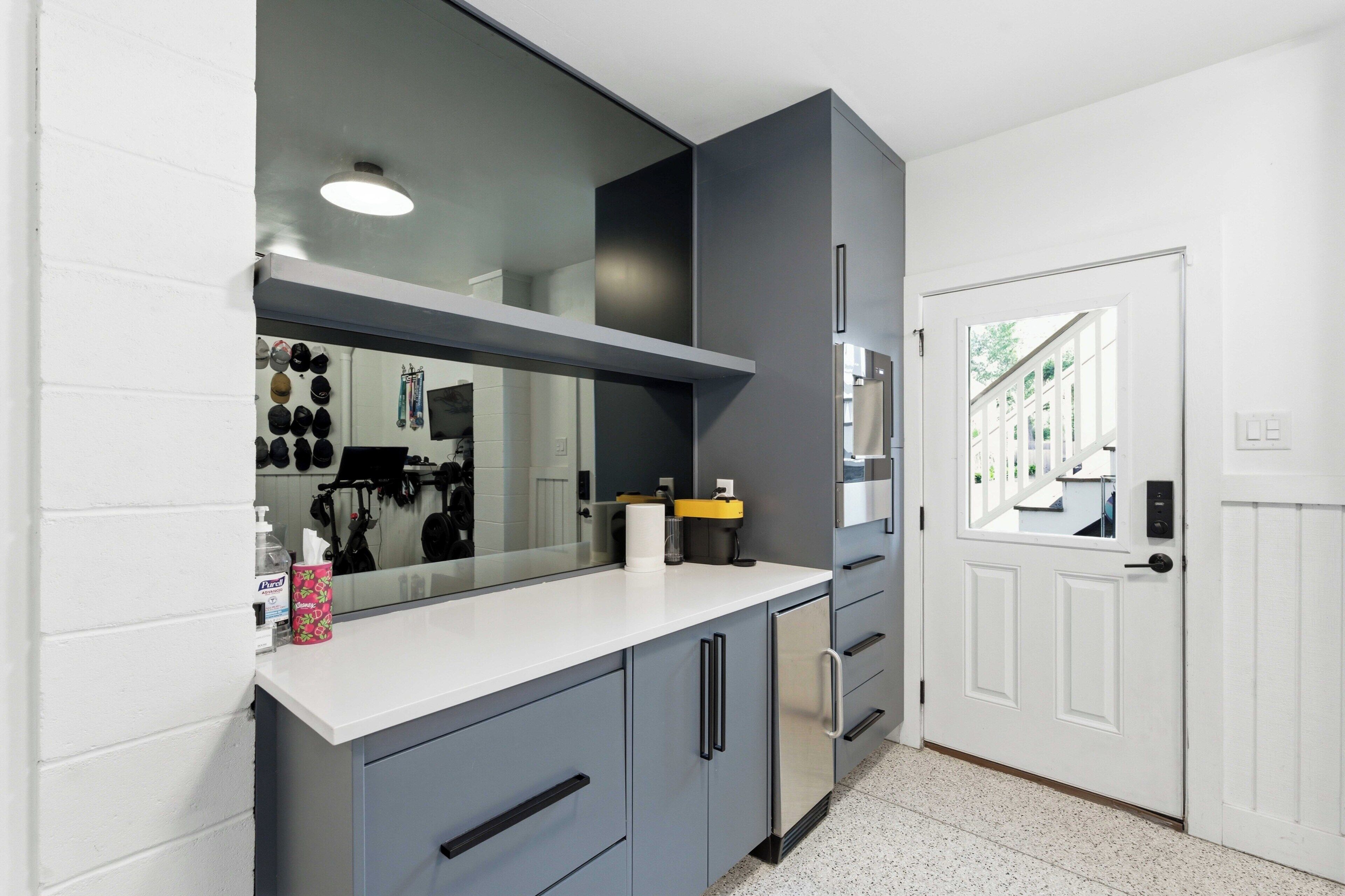 A modern kitchen with a white countertop, grey cabinets, a stainless steel refrigerator, and a large mirror above the sink.