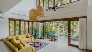 A spacious living area with a large sofa, a coffee table, and a dining table. There are large windows offering a view of the outdoors. - Tempat Santai Bali (Ubud)