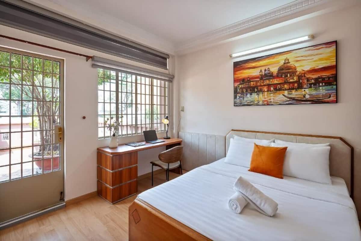 Een hotelkamer met een bed, een bureau met een laptop, een stoel, een raam met uitzicht op een gebouw en een schilderij aan de muur.