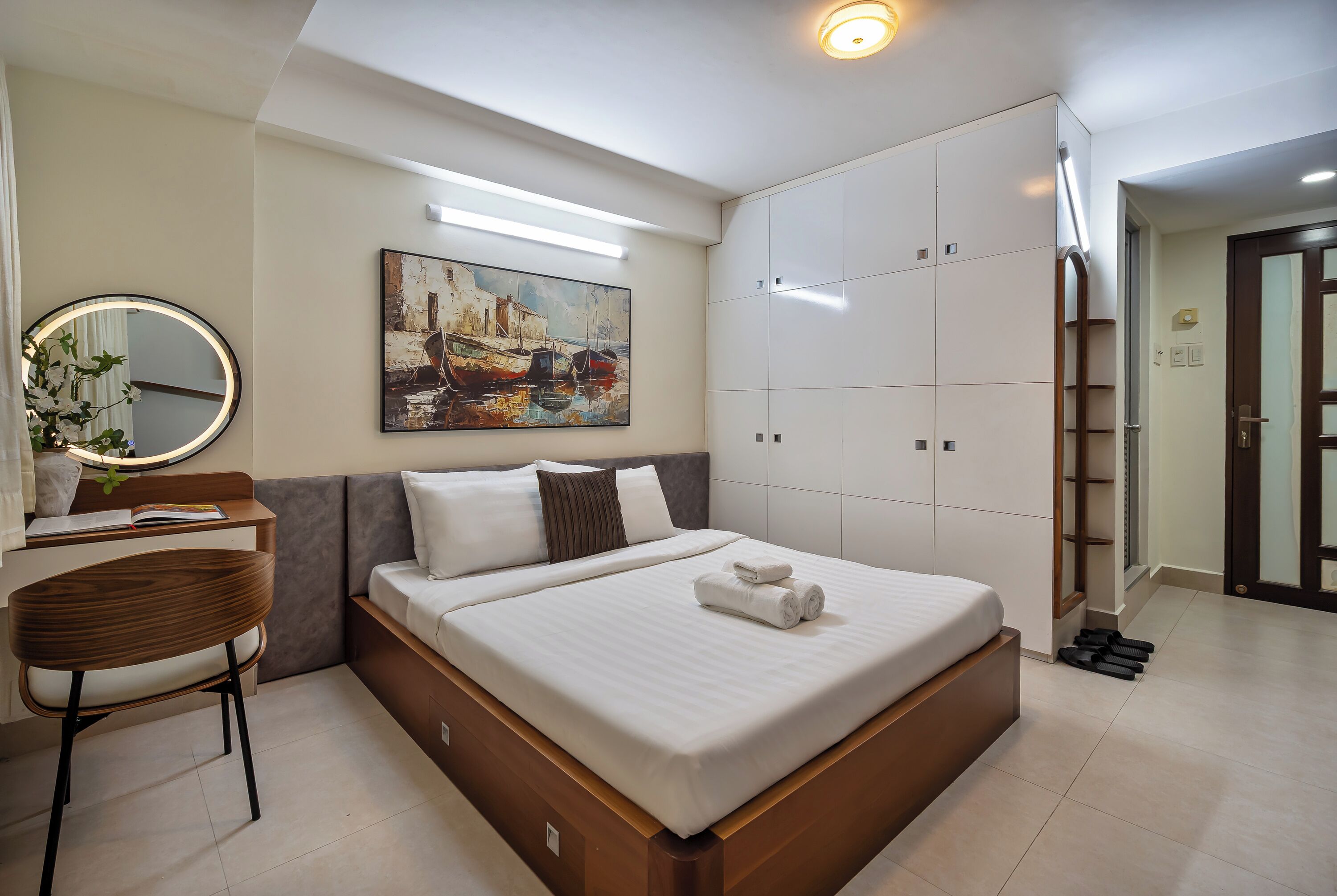 A modern bedroom with a large bed, a desk with a chair, a round mirror, and a painting on the wall.