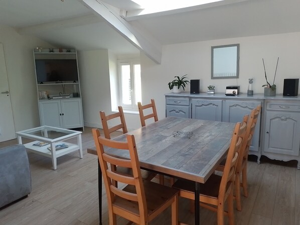 A dining area with a wooden table and chairs, a white cabinet with a TV, and a kitchen area with cabinets and a mirror. - Le nid de la clairière, renovated country house in a quiet, wooded hamlet (Heric)