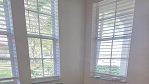 Una habitación con una mesa de madera y cuatro sillas, ventanas amplias con persianas y un enchufe visible en la pared.