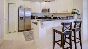 Una cocina con armarios blancos, un refrigerador de acero inoxidable, un microondas y una barra con dos taburetes.