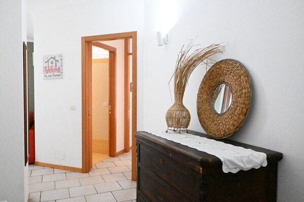 A hallway with a wooden door, a welcome sign, a wicker mirror, and a wicker vase on a dresser.