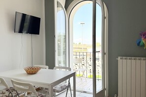 A room with a TV mounted on the wall, a dining table with chairs, a basket, and a view through a glass door to an outdoor balcony.