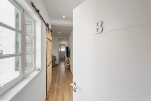 Ein Flur mit Holztür, einem Fenster mit Jalousien, einer Tür mit Beschriftung 'B' und einem Fernseher an der Wand.