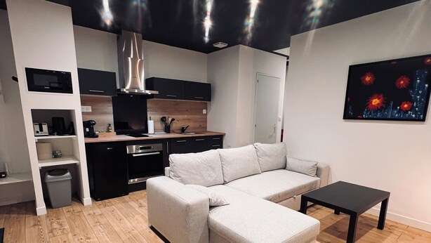 Ein modernes Wohnzimmer mit einem Ecksofa, einem kleinen schwarzen Tisch und einer Küchenzeile mit Mikrowelle und Schränken.