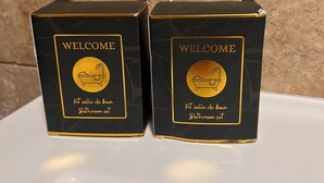 Zwei schwarze Boxen mit goldener Schrift und Symbolen, beschriftet mit 'Welcome' und 'Kit salle de bain Bathroom set', auf einer weißen Oberfläche vor einer strukturierten Wand.