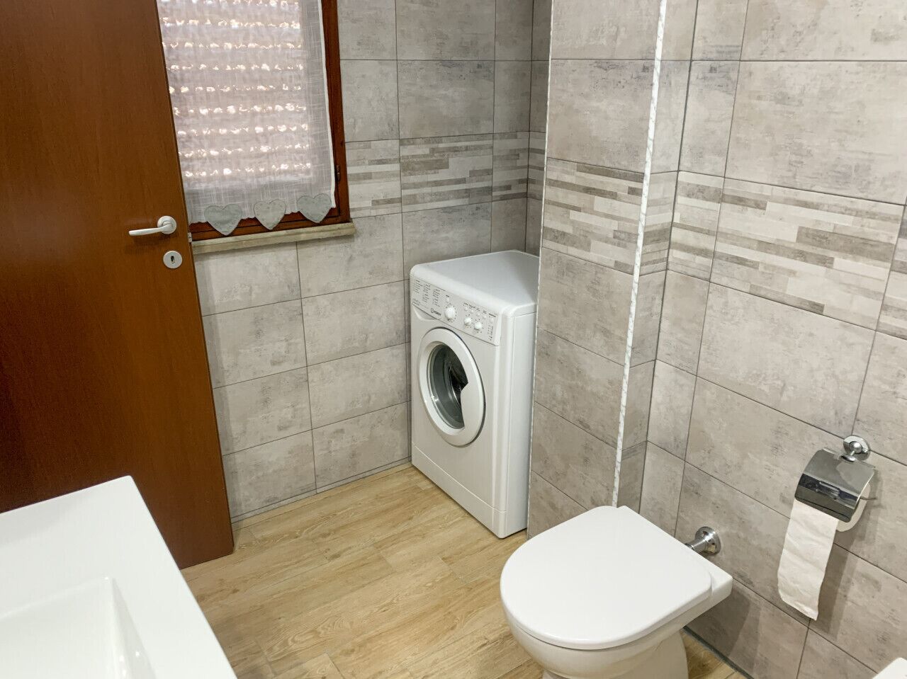 A compact bathroom with a washing machine, toilet, and a towel dispenser.