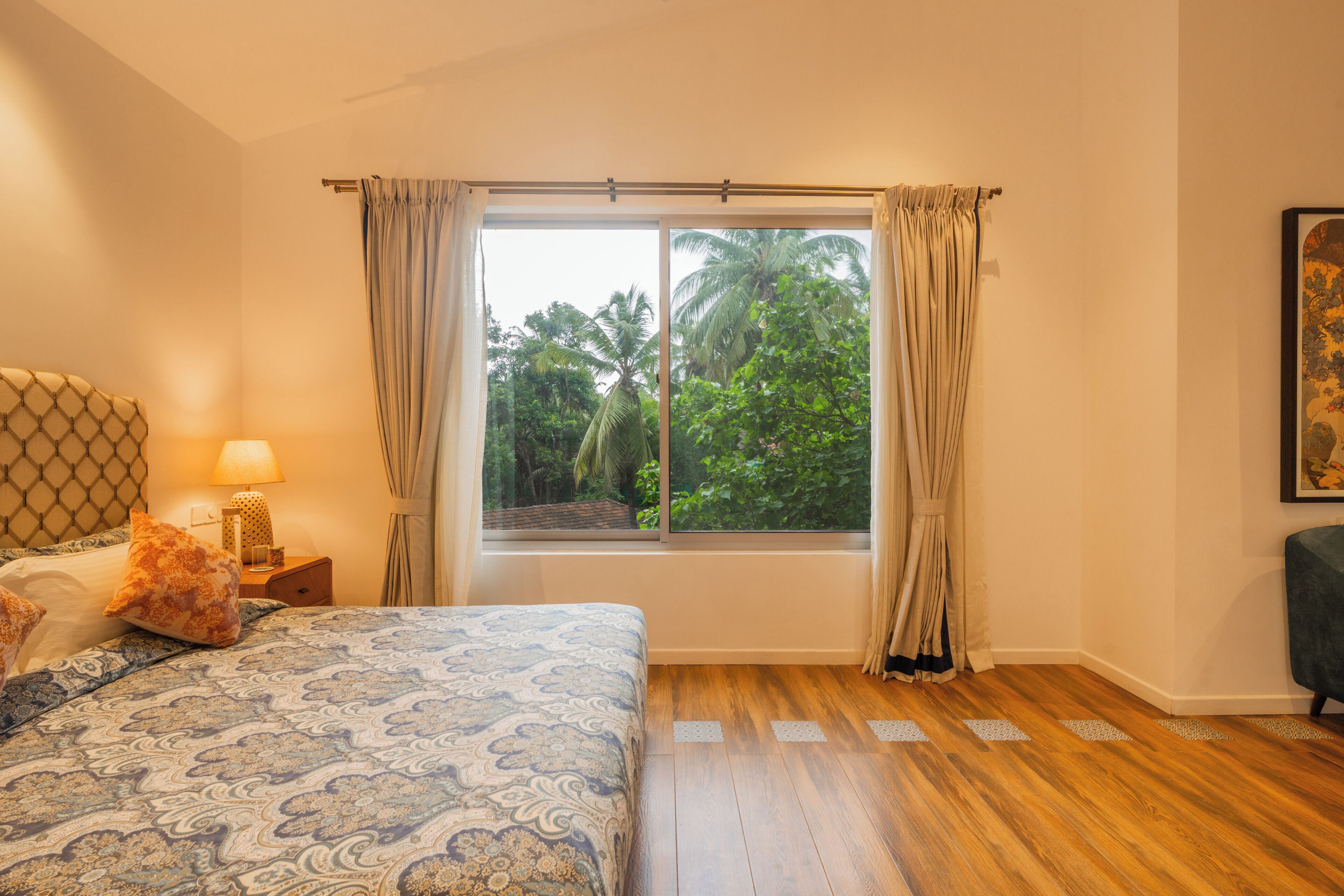 Un dormitorio con un ventanal grande, suelo de madera, una cama con ropa de cama estampada, una mesita de noche con lámpara y vistas a la vegetación exterior.