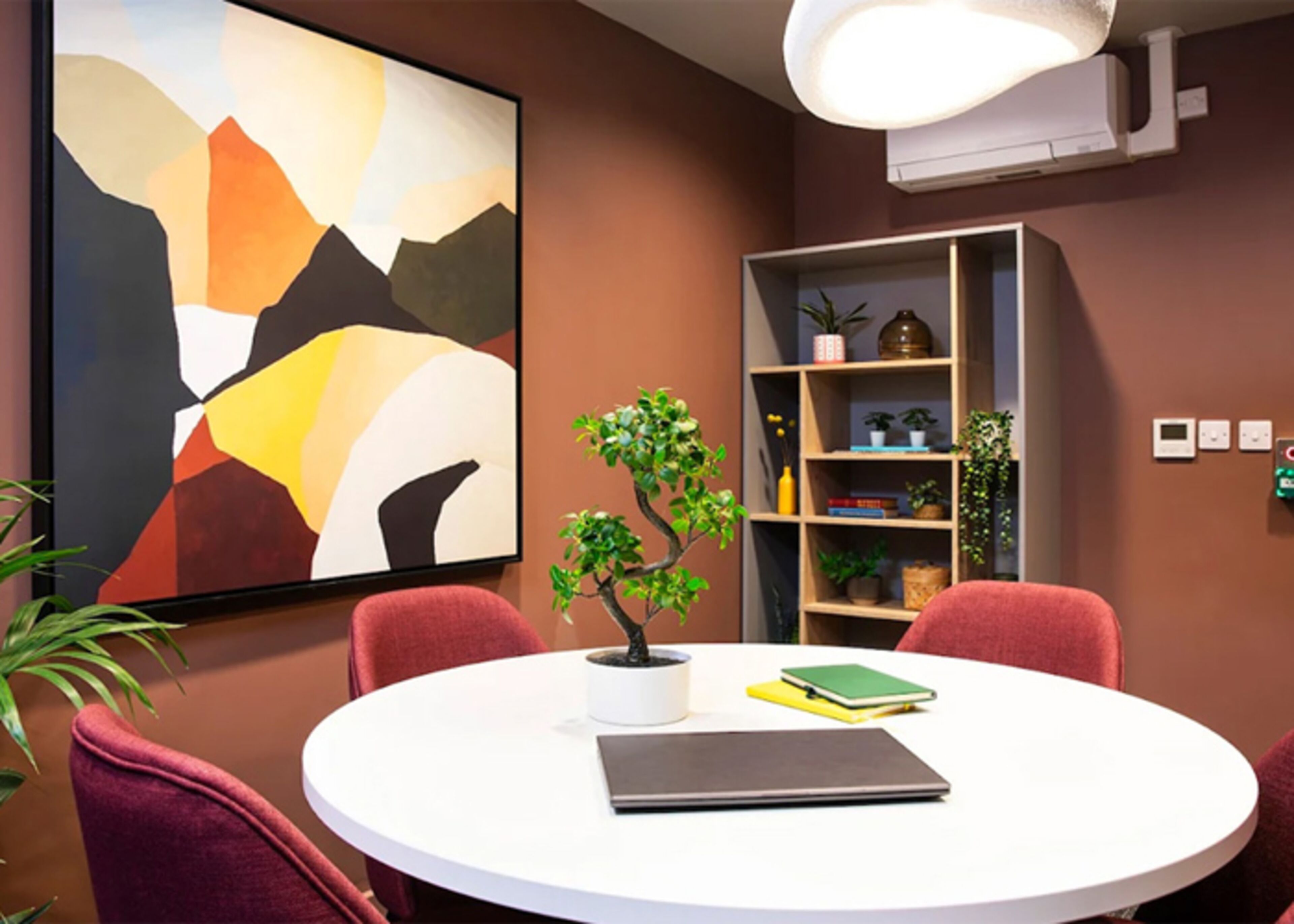 A conference room with a round table, red chairs, a potted plant, a bookshelf, and a large abstract painting on the wall.