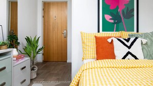 Un dormitorio con colcha a cuadros, una puerta de madera, una obra de arte enmarco y una planta en la mesita de noche.