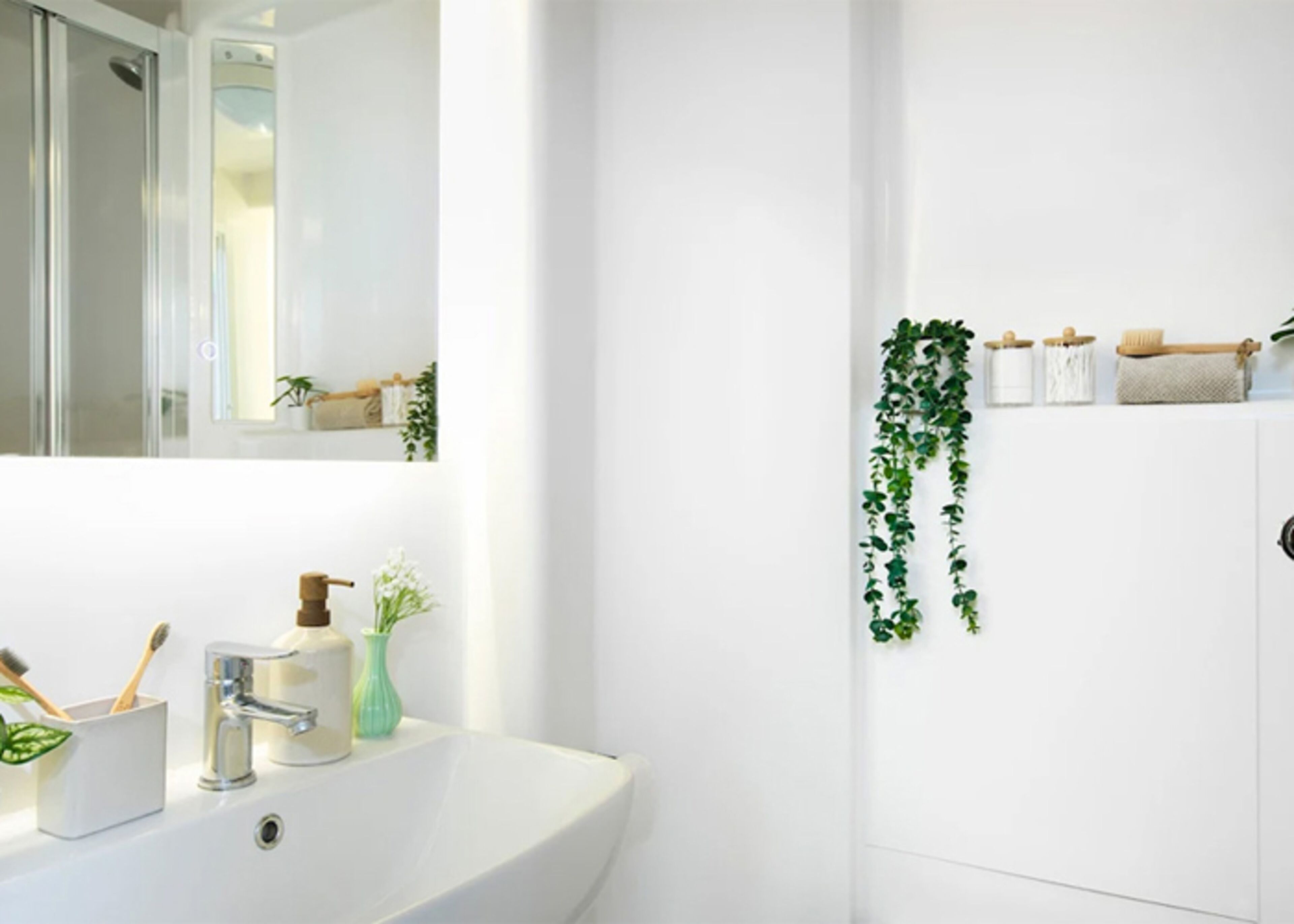 Un baño con lavamanos blanco, un espejo, ducha y estante en la pared con artículos de aseo.