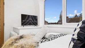 Un dormitorio con cama, televisor sobre un mueble y ventana con vista a casas.