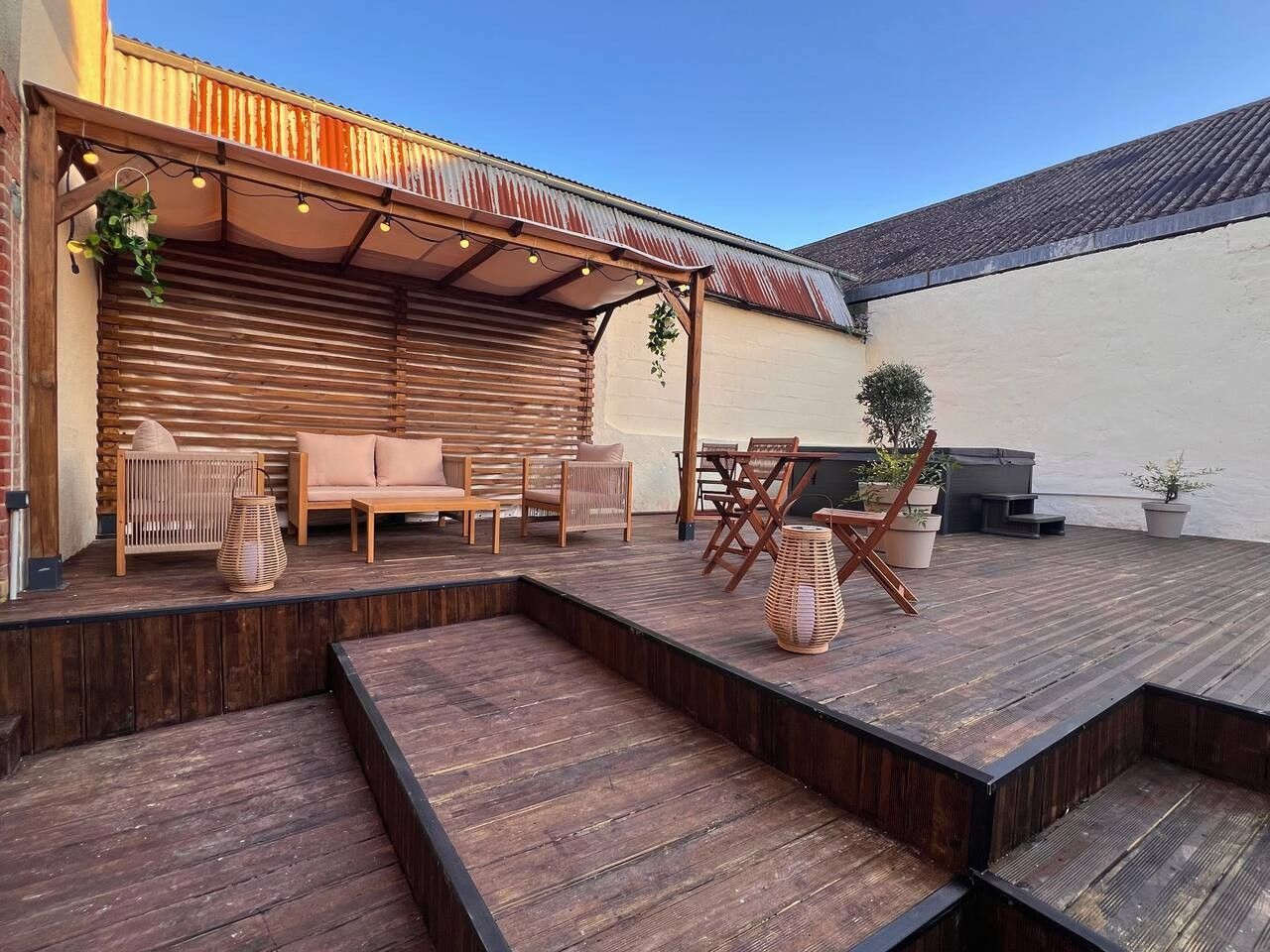 Een terras buiten met houten vloer, een overdekte zithoek en potplanten.
