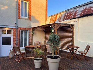 Un espace extérieur aménagé en terrasse, avec une table et des chaises en bois, des plantes en pot et une pergola décorée de guirlandes lumineuses.