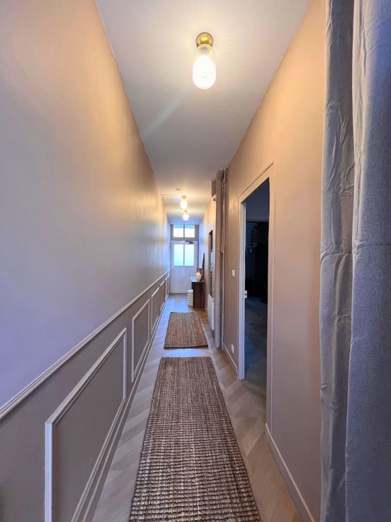 Un couloir avec un luminaire, un tapis et une porte donnant sur une autre pièce.