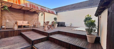 Una terraza de madera con escalones que llevan a un espacio de descanso, rodeada de macetas con plantas y una pared blanca.