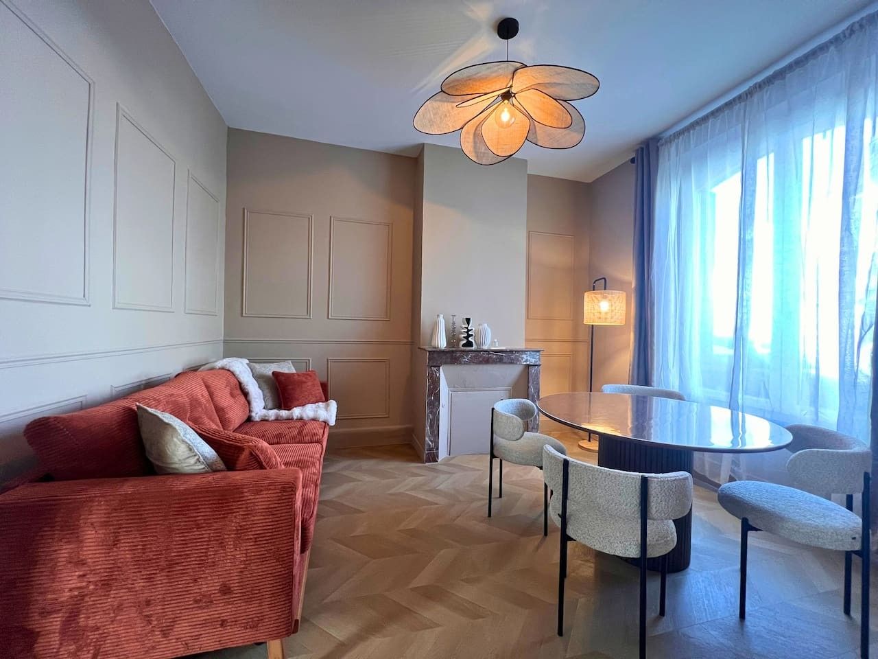 En moderne stue med sofa, spisebord med stole, pejs og et stort vindue med gardiner.