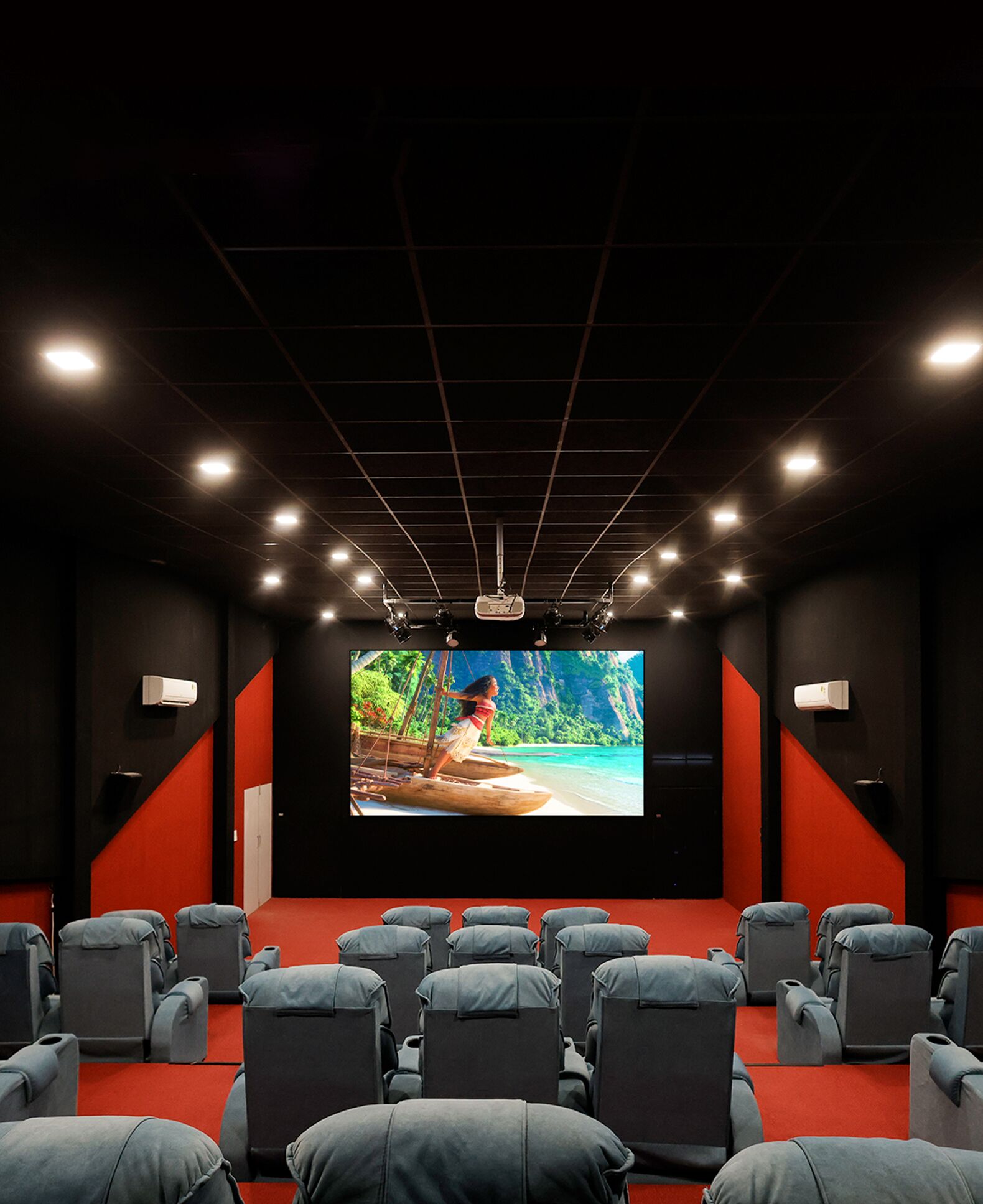 A movie theater with rows of grey seats, a large screen, and a dark ceiling with grid lighting.