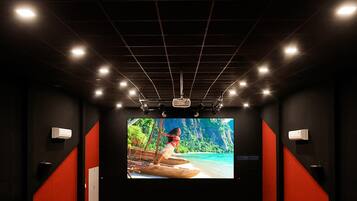 A movie theater with rows of grey seats, a large screen, and a dark ceiling with grid lighting.