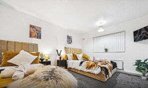 A modern bedroom with a large bed, a nightstand, a wall-mounted TV, and a window with blinds.