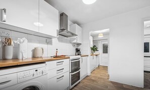 A modern kitchen with white cabinetry, a wooden countertop, a built-in washing machine, and a stainless steel range hood.