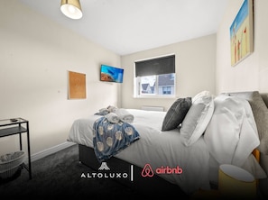 A hotel room with a bed, a desk, a chair, a window, and artwork on the walls.