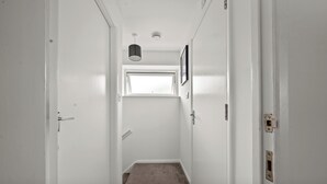 Um corredor estreito com paredes brancas, piso coberto por carpete e uma janela no final, além de uma porta à esquerda.