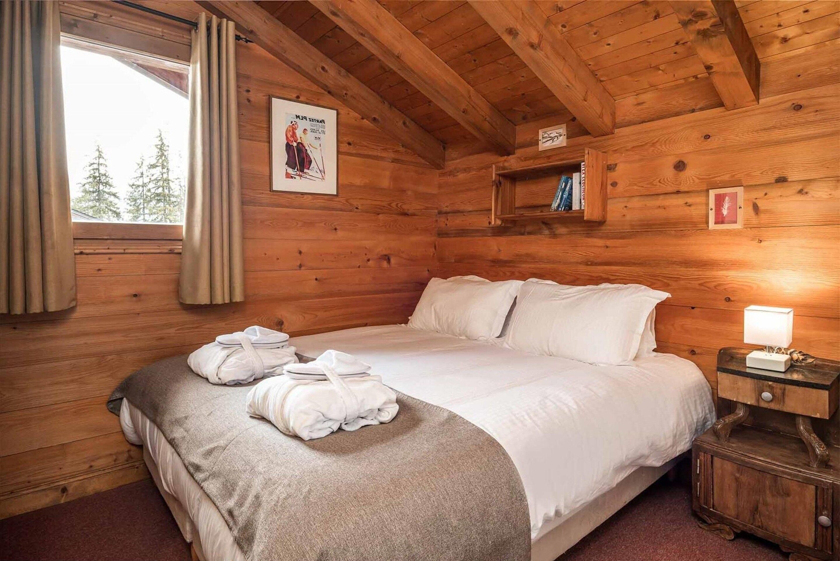 A wooden cabin room with a bed, a window with curtains, a framed picture, and a nightstand with a lamp.