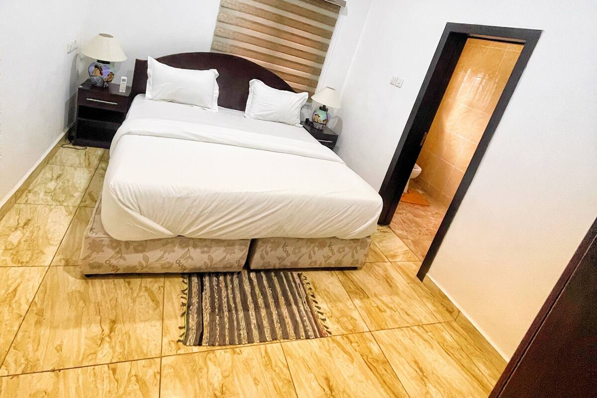 Habitación de hotel con cama, mesita de noche, lámpara, aire acondicionado y ventana con persianas.