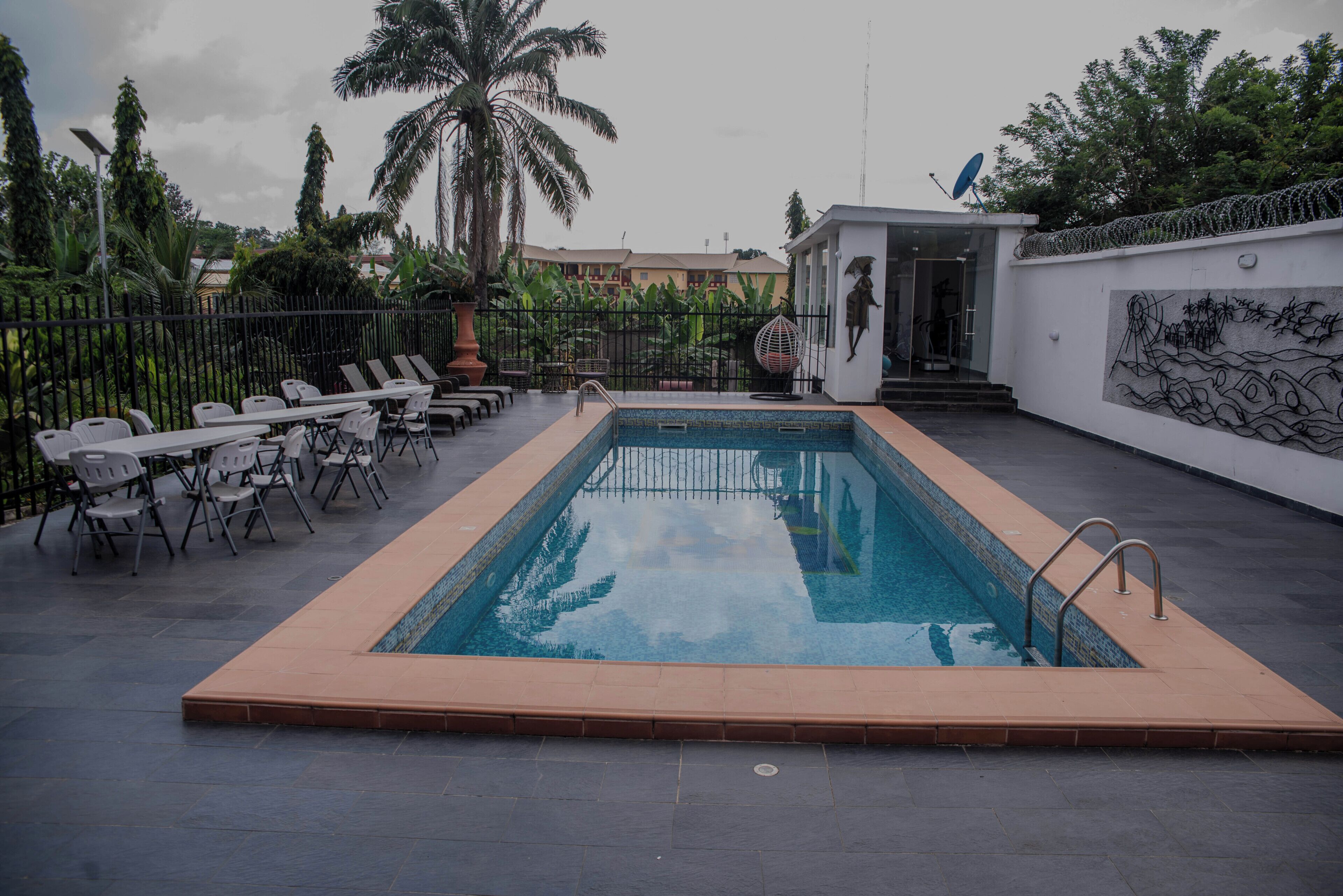 Une piscine rectangulaire bordée de carreaux, entourée de chaises longues et d’un petit bâtiment surmonté d’une antenne parabolique.
