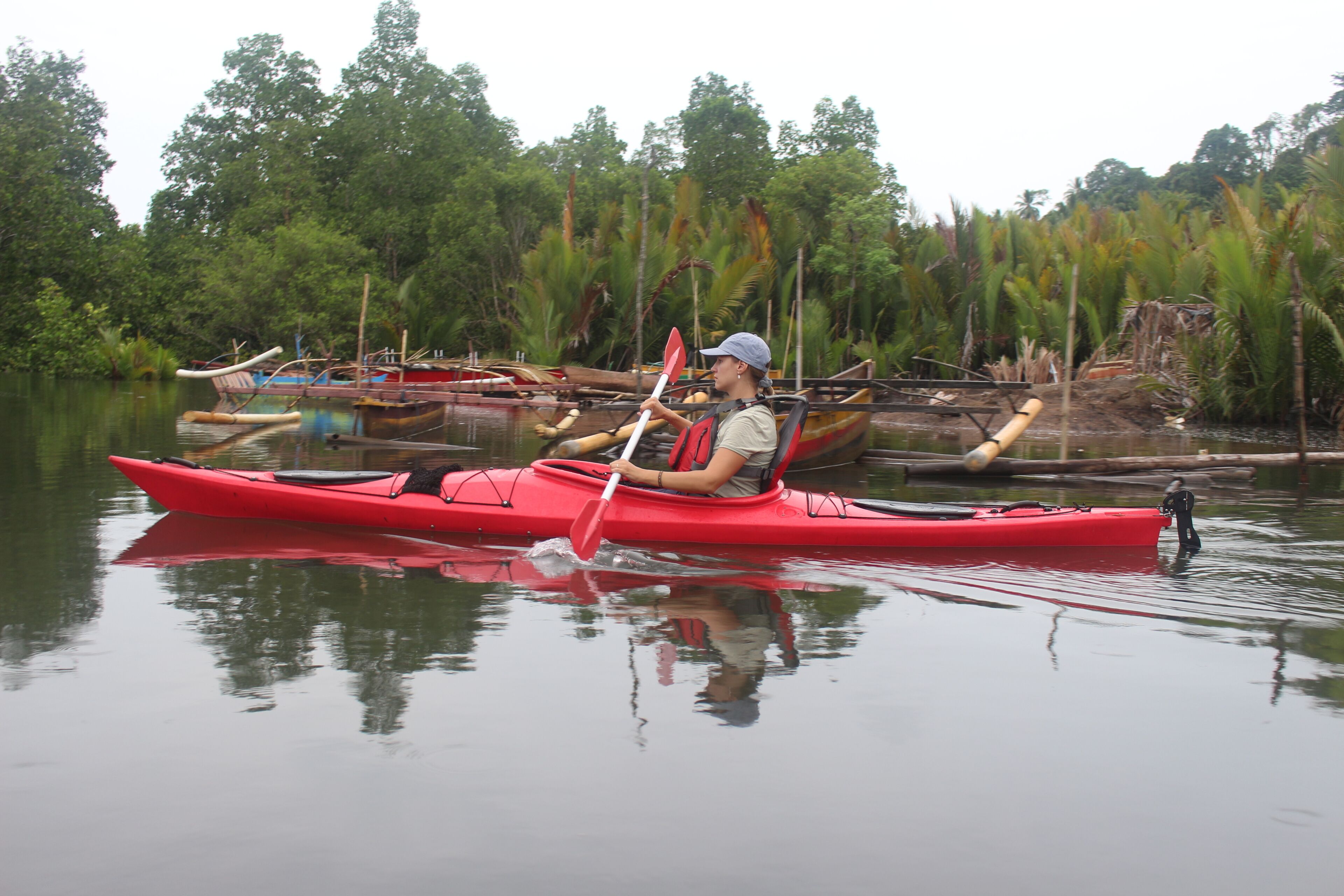 A person kayaking on calm water with boats and dense greenery in the background.