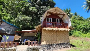 A traditional thatched-roof house with a balcony, surrounded by lush greenery and palm trees. - Pondok Wisata Noah (Likupang)
