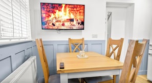 Un coin repas avec une table et des chaises en bois, un téléviseur diffusant une image de cheminée et une fenêtre avec des stores.