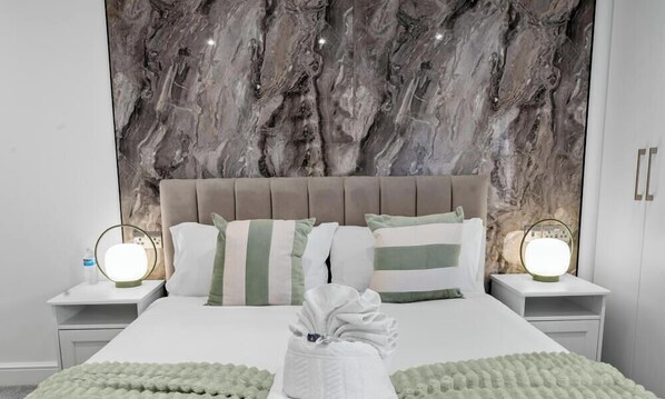Habitación de hotel con cabecera de mármol, cama con almohadas a rayas, lámparas de noche y una cama impecable con manta verde.