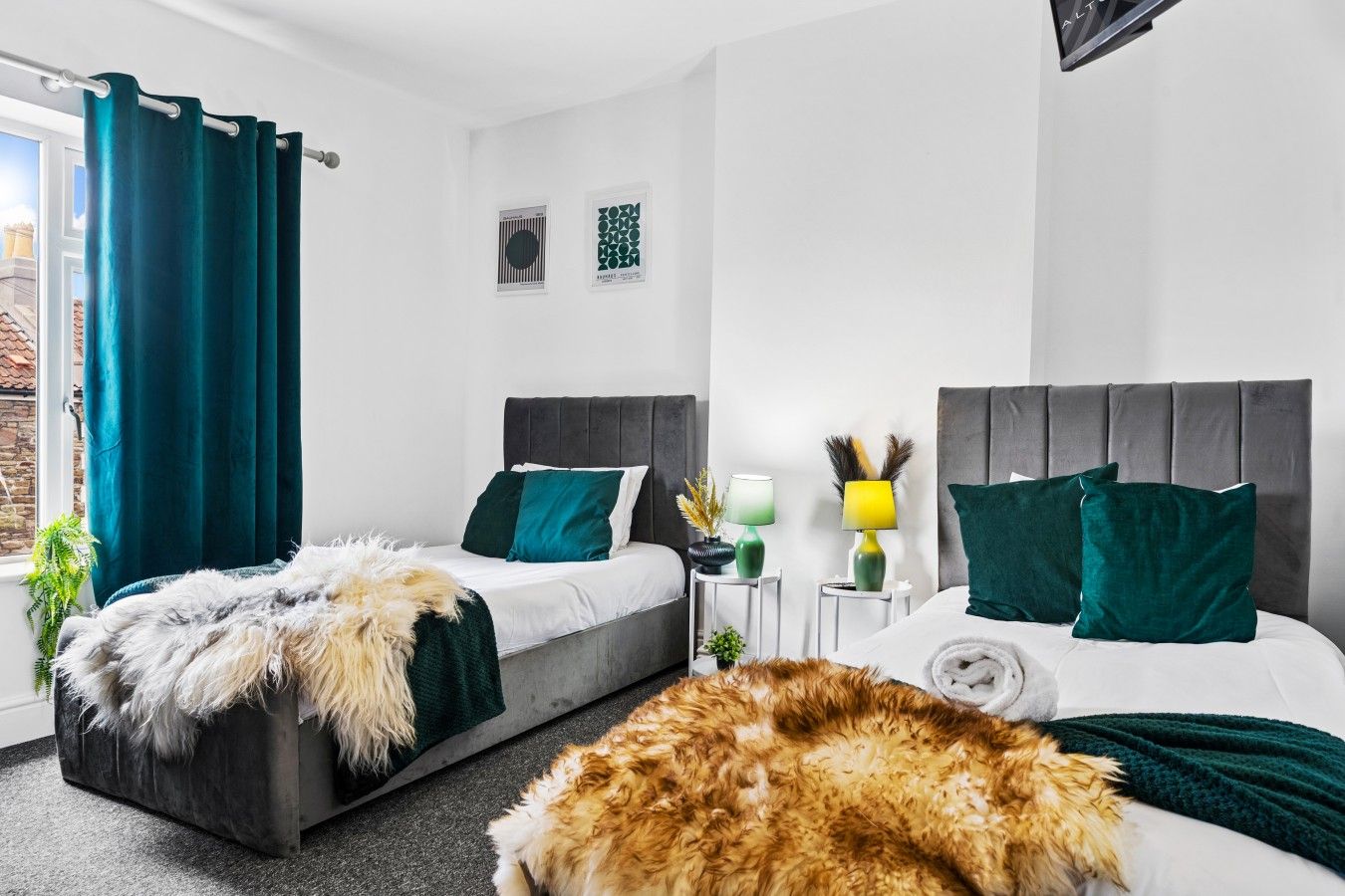 Une chambre moderne avec deux lits, une grande fenêtre avec des rideaux, une petite table de chevet avec des lampes et un plaid décoratif en fausse fourrure.